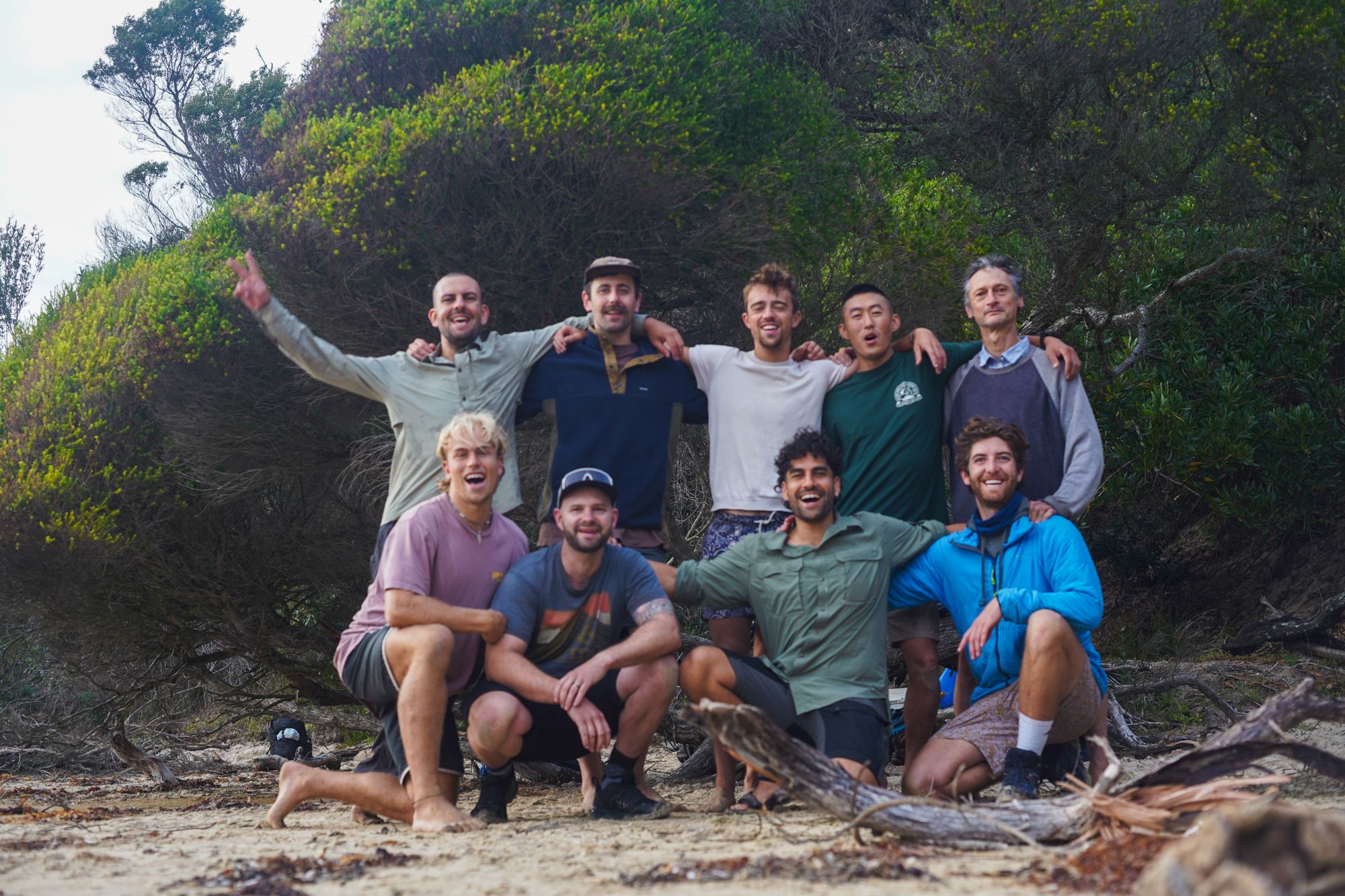 Eleven men grinning on a beach, arms round each other