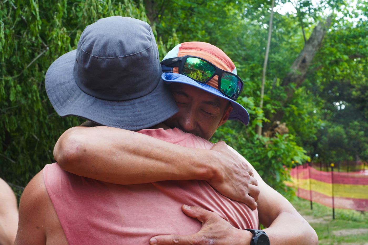 Two men hugging on a riverbank, faces hidden under sun hats
